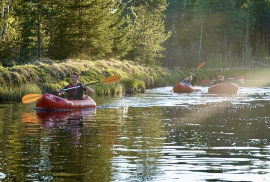 tre personer paddlar i varsin uppblåsbar packraft i solnedgången. Stilla vatten och grön skog i bakgrunden