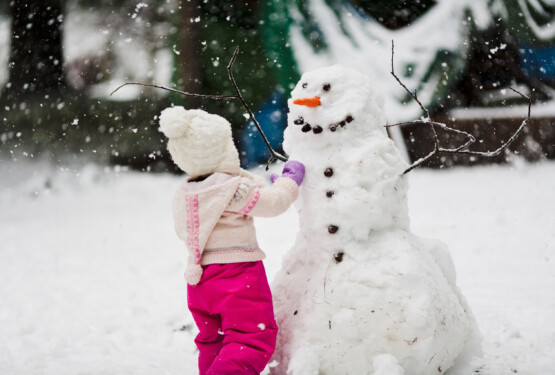 A toddler girl in a pink snowsuit with wooly hat toque builds a large snowman with carrot nose and smiling face in backyard with snowflakes snow falling in winter at Christmas
